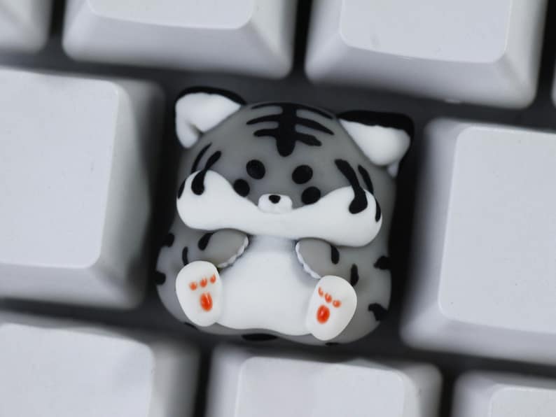 May include: A gray and black tiger-striped keycap with white paws and orange paw pads. The keycap is sitting on a white keyboard.