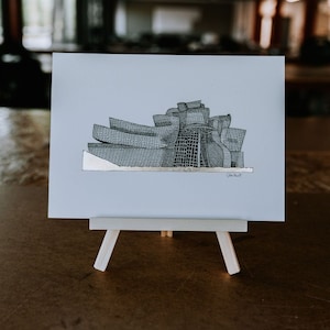 May include: A black and white line drawing of the Guggenheim Museum in Bilbao, Spain. The drawing is on a light blue background and is framed on a small easel.