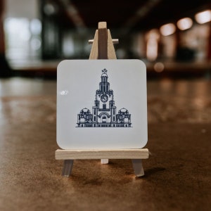 Puede incluir: Un posavasos blanco con una ilustración en blanco y negro de un edificio con una torre del reloj. El posavasos se muestra en un pequeño caballete de madera.