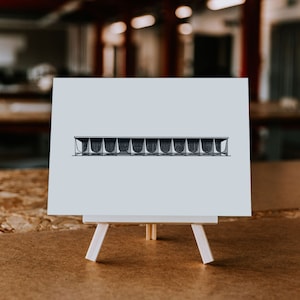 May include: A black and white line drawing of a modern building with a curved roof, printed on a white card and displayed on a small easel.