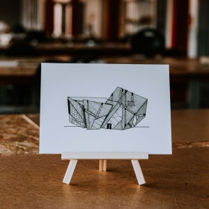 May include: A black and white line drawing of a modern building with a geometric design. The drawing is on a white card mounted on a small wooden easel.