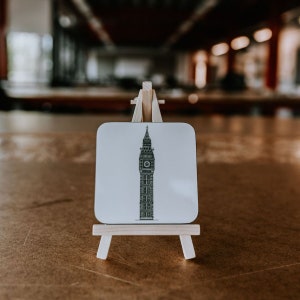 Könnte beinhalten: Ein weißer Untersetzer mit einer Schwarzweiß-Illustration des Big Ben auf einem kleinen Holzständer.