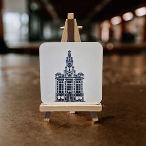 Puede incluir: Un posavasos blanco con un dibujo en blanco y negro de un edificio con una torre del reloj. El posavasos está en un pequeño caballete de madera.