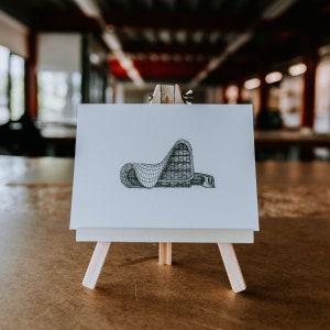 May include: A black and white line drawing of a modern building on a small white easel. The drawing is on a white card and the easel is made of wood.