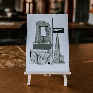 May include: A white card with black line drawings of three modern buildings. The card reads "Architects have to dream..." and is displayed on a small wooden easel.