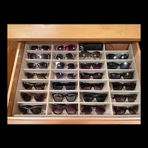 May include: A wooden drawer filled with various sunglasses. The sunglasses are arranged in individual compartments, showcasing a variety of frame styles and lens colors, including black, brown, and tortoiseshell.