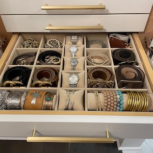 May include: A white drawer with a gold handle, open to reveal a beige velvet-lined organizer with compartments for storing jewelry and accessories. The organizer contains a variety of belts, watches, bracelets, and bangles.