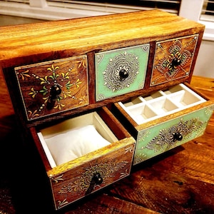 May include: Wooden jewelry box with three drawers, two open. The drawers are decorated with intricate floral and geometric patterns in shades of brown, green, and gold. The top of the box is a natural wood color, and the drawers have black knobs.