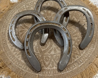Può includere: Quattro ferri di cavallo argentati e usurati disposti su una superficie di juta beige strutturata. I ferri di cavallo sono curvi, con fori per i chiodi, e probabilmente in metallo. Suggerisce un tema rustico o western.