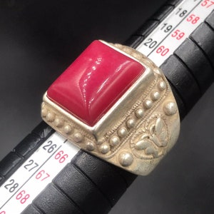 May include: A silver ring featuring a large, square, red gemstone set in a bezel setting. The ring band is decorated with a floral design and a row of small beads.