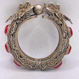 Puede incluir: Un brazalete de plata con intrincados diseños de dragones y acentos de piedras preciosas rojas. El brazalete tiene bisagras y se abre para ajustarse a la muñeca.
