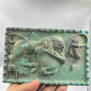 Könnte beinhalten: Eine rechteckige Bronzeplatte mit einem Relief von drei Löwen. Die Platte hat eine grüne Patina und einen perlenbesetzten Rand.