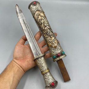 May include: An antique dagger with a silver handle and sheath. The handle and sheath are adorned with intricate patterns and red and turquoise stones. The blade is silver and the sheath is made of bone.