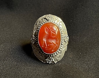 Könnte beinhalten: Ein silberner Ring mit einem ovalen Karneol-Stein, der in der Mitte eingesetzt ist. Der Stein ist mit einem detaillierten Design einer mythischen Kreatur verziert. Der Ring ist mit komplizierten floralen Mustern verziert.