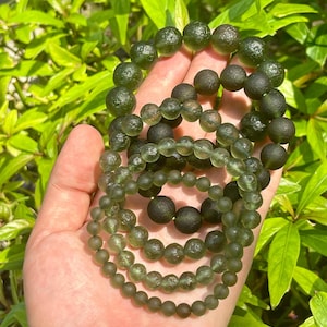 May include: Several green beaded bracelets of varying sizes are displayed. The beads have a translucent quality, with a range of green hues. The bracelets are held against a backdrop of green foliage, creating a natural aesthetic.
