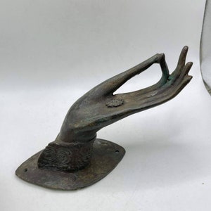 May include: A bronze door handle shaped like a hand in a gesture of giving. The hand is detailed with a bracelet and a ring. The handle is mounted on a rectangular plate.