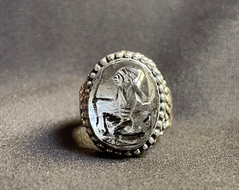 Könnte beinhalten: Ein silberner Ring mit einem klaren Stein, der ein geschnitztes Design einer mythischen Kreatur zeigt. Der Stein ist in einer silbernen ovalen Fassung mit einem perlenbesetzten Rand gefasst.