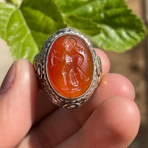 May include: A silver ring with an orange carnelian stone featuring an engraved image of a warrior holding a sword and shield.