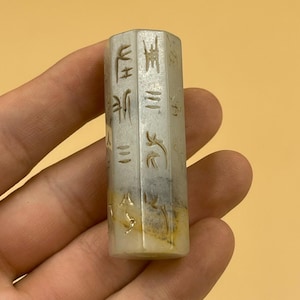 May include: A white jade cylinder with Chinese characters carved into the sides. The cylinder is approximately 2 inches long and 1 inch in diameter.