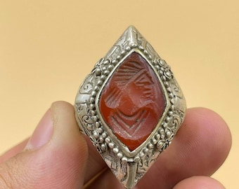 Könnte beinhalten: Ein silberner Ring mit einem filigranen Design und einem roten Karneol-Stein, der in der Mitte eingesetzt ist. Der Stein hat ein geschnitztes Design.