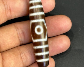 May include: A brown and white dzi bead with a cylindrical shape. The bead features a central eye design and horizontal stripes. The bead is held in a hand.