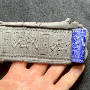 May include: A gray clay tablet with carved figures of animals and symbols, next to a blue lapis lazuli cylinder seal. The tablet features a series of stylized animals and rectangular panels. The cylinder seal is a deep blue color with white veining.
