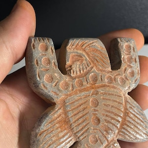 Puede incluir: Una figurita de piedra tallada de una persona con un tocado y detalles intrincados. La figurita es marrón y gris con una superficie texturizada.