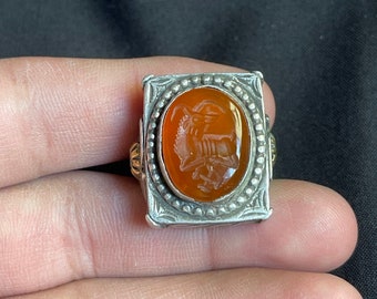 Könnte beinhalten: Rechteckiger Silberring mit einem ovalen, orangefarbenen Stein. Der Stein zeigt ein detailliertes Ziegenmotiv. Der Ring hat goldfarbene Akzente und einen dekorativen Rand.