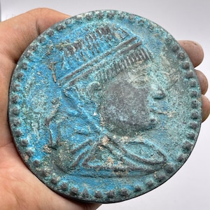 Könnte beinhalten: Eine runde Bronzemünze mit einem detaillierten Profil einer Person, die eine Krone oder einen Kopfschmuck trägt. Die Münze hat eine blaugrüne Patina und einen perlenbesetzten Rand.