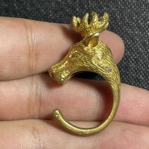 Pode incluir: Um anel de cabeça de alce dourado com acabamento texturizado. O anel é aberto e tem um gancho na lateral.