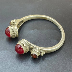 Puede incluir: Un brazalete de plata con tres acentos de piedras preciosas rojas. El brazalete está abierto y tiene un diseño decorativo.