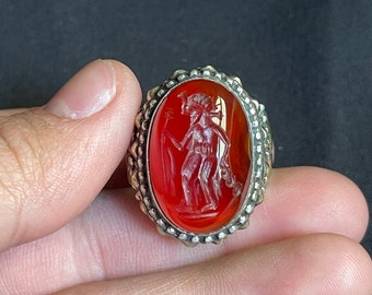 Könnte beinhalten: Ovale Ring mit rotem Stein, der eine geschnitzte Figur mit Stab zeigt. Der Ring hat ein silberfarbenes, strukturiertes Band. Die Figur ist detailliert mit Linien und Schatten, vor einem leuchtend roten Hintergrund.