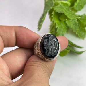 Puede incluir: Un anillo de plata con una piedra ovalada negra que presenta un diseño grabado de una persona sosteniendo un bastón.