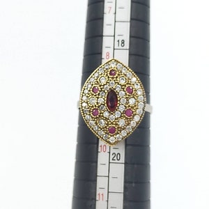 May include: A silver ring with a gold-toned center featuring a large red gemstone surrounded by smaller white and red gemstones. The ring is sized on a ring sizer.