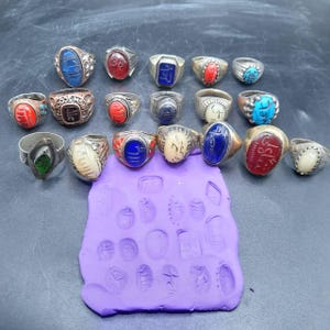 May include: A collection of 18 silver rings with various colored stones and engraved designs. The rings are arranged on a purple surface with impressions of the rings' designs.