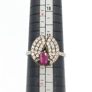 Könnte beinhalten: Ein silberner Ring mit einem roten Edelstein, der von klaren Steinen umgeben ist. Der Ring befindet sich auf einem Lineal mit Markierungen von 8 bis 21.