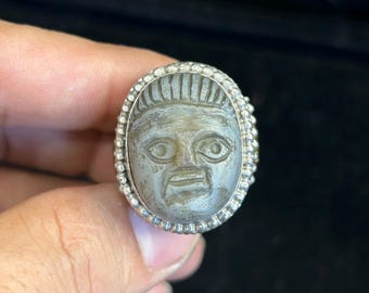 Könnte beinhalten: Ovaler Ring mit einem geschnitzten Gesichtsdesign. Das graue Gesicht weist detaillierte Merkmale auf und ist in einem silberfarbenen Band eingefasst. Das Band ist mit kleinen, runden Akzenten um das Gesicht verziert.