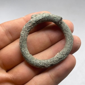 May include: A weathered, circular metal object with a textured surface and a greenish-gray hue. The ring-shaped item has a raised, decorative element at the top. The object is held in a hand, suggesting its size and scale.