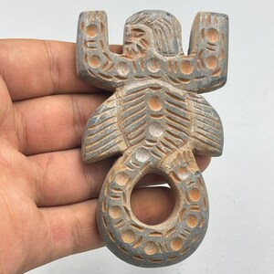 Puede incluir: Una figurita de piedra tallada de una criatura mítica con un anillo en la cola. La criatura tiene un cuerpo humanoide con alas y una cola que se curva en un anillo. La piedra es de color marrón claro con una pátina gris.