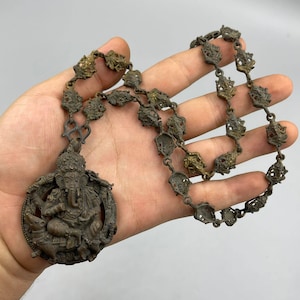 May include: A brass necklace with a detailed pendant featuring the Hindu deity Ganesha. The pendant is circular and depicts Ganesha sitting on his mount, a mouse, with his trunk raised. The necklace is made of a chain of intricately designed links.