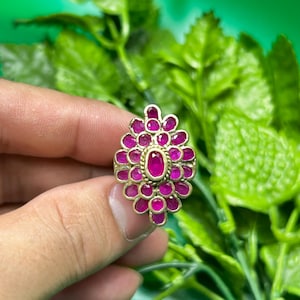 Puede incluir: Un anillo dorado con un diseño floral, engastado con numerosas piedras preciosas pequeñas, redondas y de color magenta alrededor de una piedra central ovalada magenta. El anillo se presenta sobre un fondo de hojas verdes.