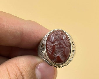 Könnte beinhalten: Ein silberner Ring mit einem roten Karneol-Stein, der mit einem gravierten Design verziert ist. Der Stein ist oval geformt und in einem silbernen Band mit filigranen Details gefasst.