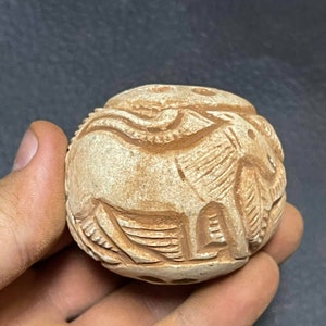 Può includere: Una sfera di pietra rotonda e scolpita con una rappresentazione dettagliata di un elefante. La sfera è di colore marrone chiaro e ha una superficie liscia e lucida.