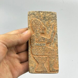 Puede incluir: Una talla rectangular de piedra de una figura sentada que lleva un tocado. La figura está tallada en relieve y tiene un diseño detallado. La piedra es de color marrón claro con una textura rugosa.