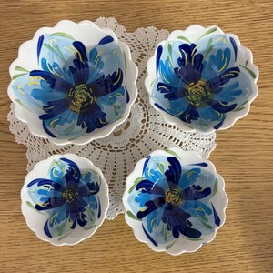 Puede incluir: Juego de cuatro cuencos de cer&aacute;mica blanca con borde festoneado. Cada cuenco presenta un dise&ntilde;o floral azul pintado a mano con detalles amarillos y verdes. Los cuencos est&aacute;n dispuestos sobre un tapete de encaje blanco, ideal para servir aperitivos.