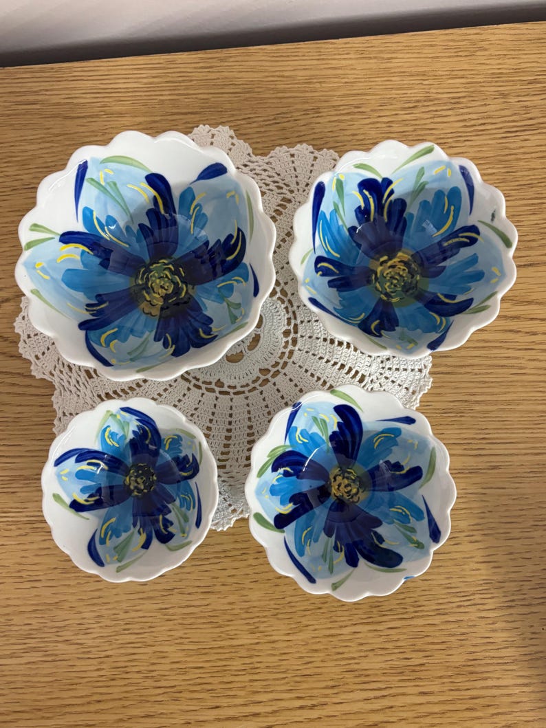 Puede incluir: Juego de cuatro cuencos de cer&aacute;mica blanca con borde festoneado. Cada cuenco presenta un dise&ntilde;o floral azul pintado a mano con detalles amarillos y verdes. Los cuencos est&aacute;n dispuestos sobre un tapete de encaje blanco, ideal para servir aperitivos.