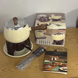 May include: A cream-colored fondue pot with a brown base, a silver lid, and a wooden handle. The set includes a box labeled 2 QT. (1.9L) DELUXE FONDUE WITH SIX FORKS, a plastic-wrapped set of six metal forks, and an instruction booklet titled IT'S FUN TO FONDUE.