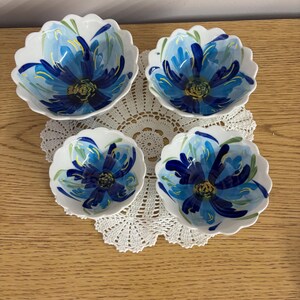 Puede incluir: Cuatro cuencos de cer&aacute;mica blanca con bordes festoneados, cada uno adornado con un vibrante dise&ntilde;o floral azul. Las flores tienen un centro azul oscuro y p&eacute;talos azul claro, con detalles verdes. Los cuencos est&aacute;n colocados sobre un tapete de encaje blanco.