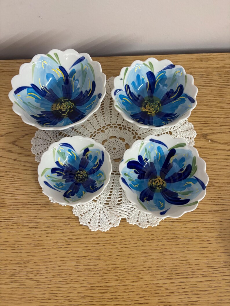 Puede incluir: Cuatro cuencos de cer&aacute;mica blanca con bordes festoneados, cada uno adornado con un vibrante dise&ntilde;o floral azul. Las flores tienen un centro azul oscuro y p&eacute;talos azul claro, con detalles verdes. Los cuencos est&aacute;n colocados sobre un tapete de encaje blanco.