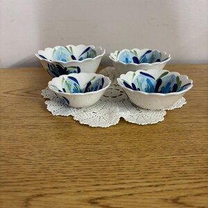 Puede incluir: Cuatro cuencos de cer&aacute;mica blanca con bordes festoneados, cada uno adornado con un dise&ntilde;o floral azul. Los cuencos est&aacute;n dispuestos sobre un tapete de encaje blanco, sobre una superficie de madera clara. El patr&oacute;n floral presenta tonos de azul y verde.
